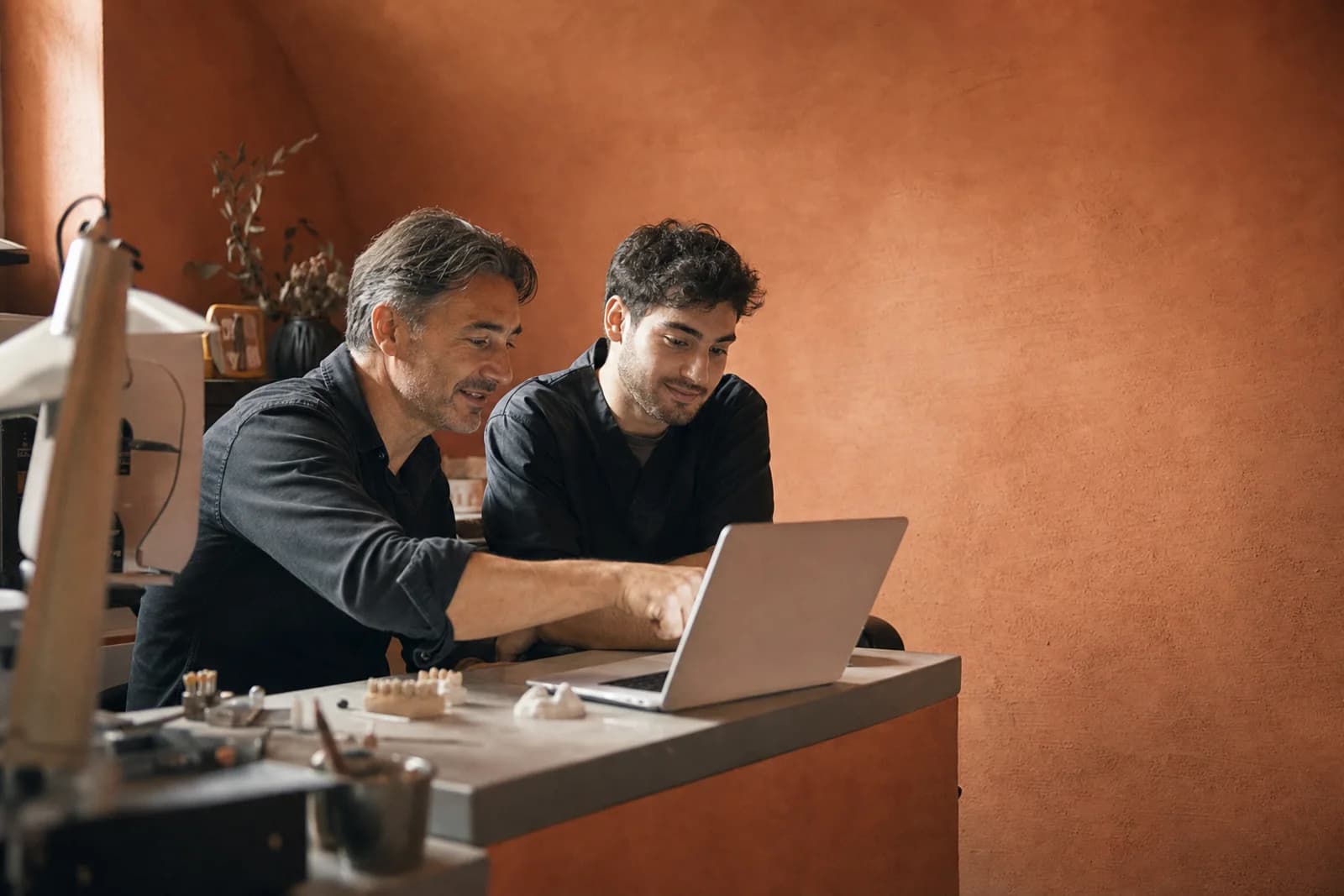 Meister und Azubi sitzen nebeneinander am Schreibtisch vor einem Laptop, der Meister zeigt auf den Bildschirm